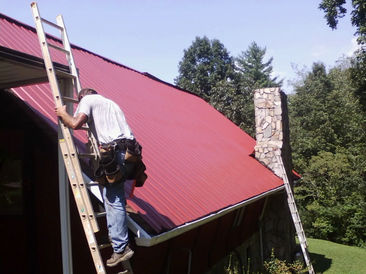 Roofer on a ladder beside a red metal roof and stone chimney — Soffit & Fascia Repair by Roofing Brothers in Stonewall